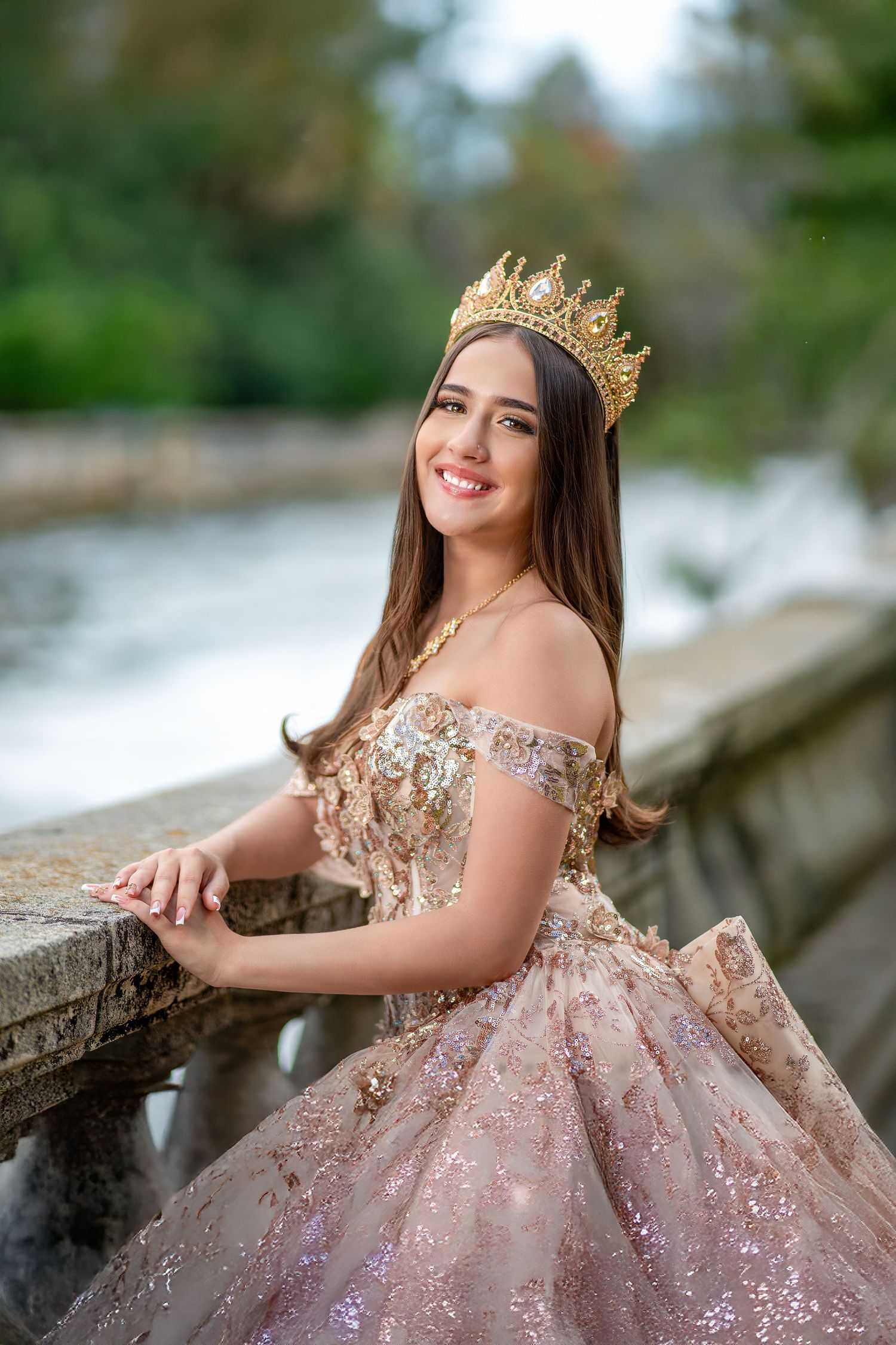 A person in a sparkly off-shoulder pink ball gown and gold crown poses by a stone railing near a waterfall.