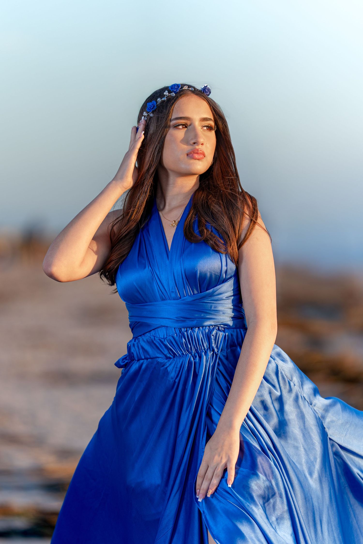 A person in a flowing royal blue dress and floral headpiece poses outdoors at sunset.