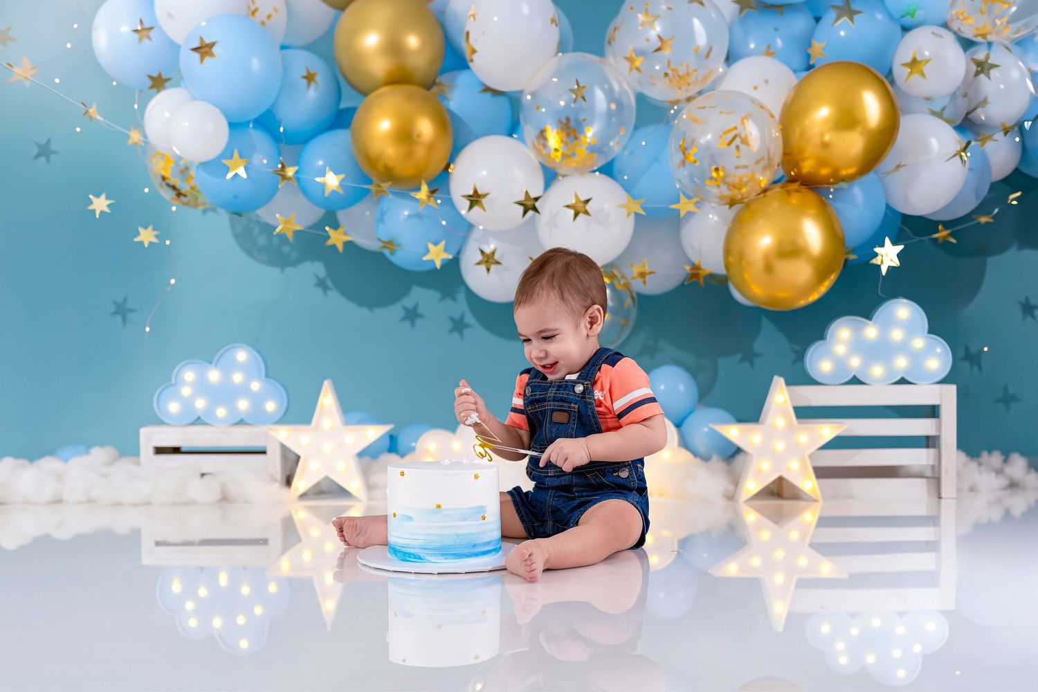 Baby boy in denim overalls smashing first birthday cake surrounded by blue, white, and gold balloons and star decorations.