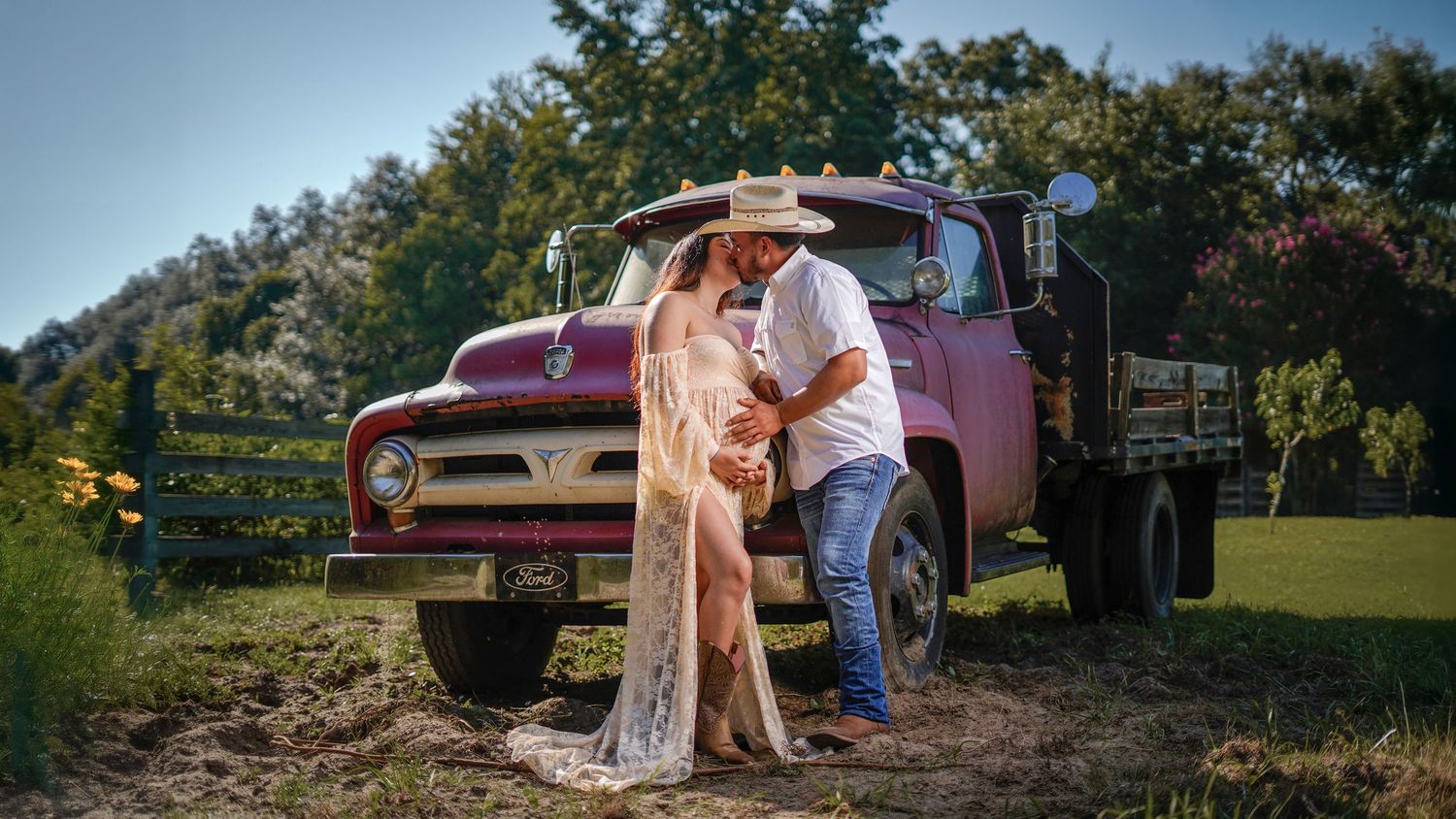 Couple in western wear standing beside a vintage pink Ford flatbed truck in a rustic outdoor setting at sunset.