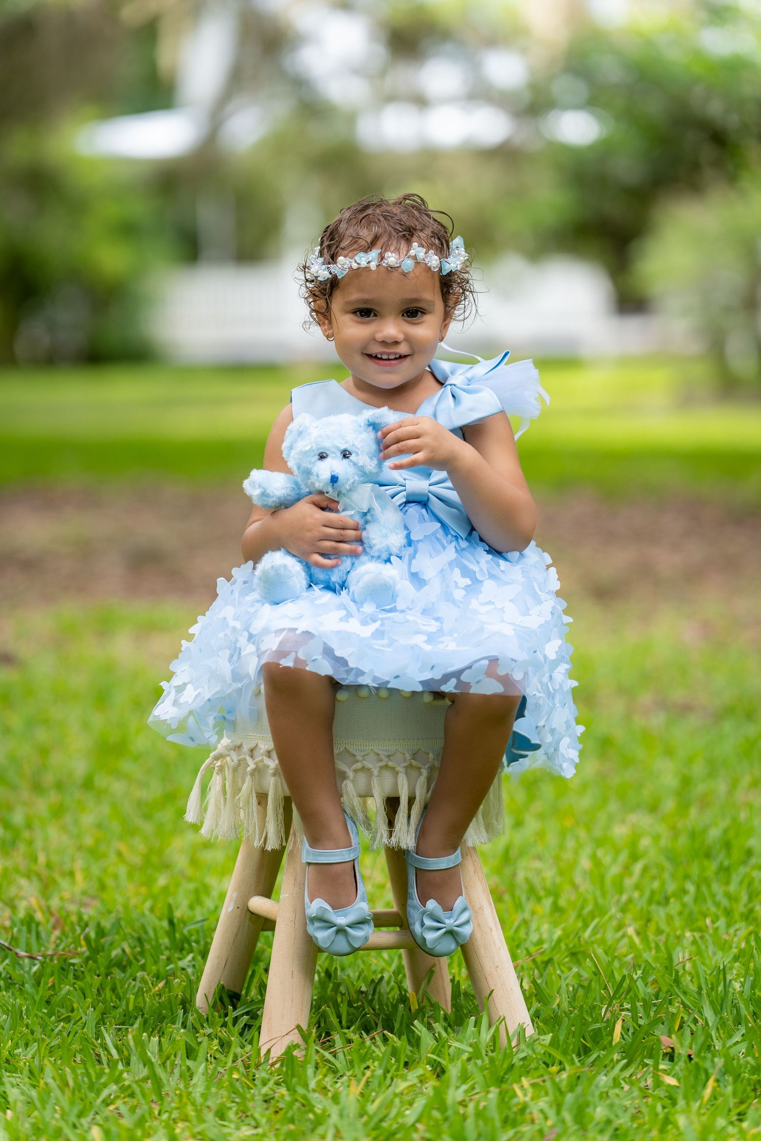 A small child sits on a wooden bench outdoors while holding a blue teddy bear on a sunny afternoon.