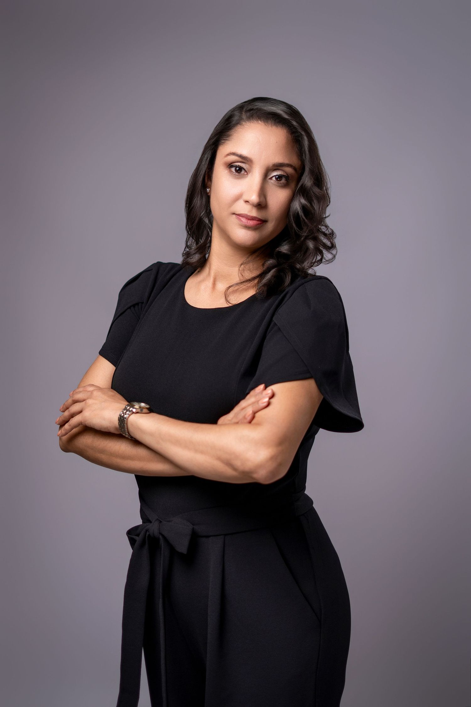 Professional woman in black outfit with arms crossed, posing confidently against a gray studio background.