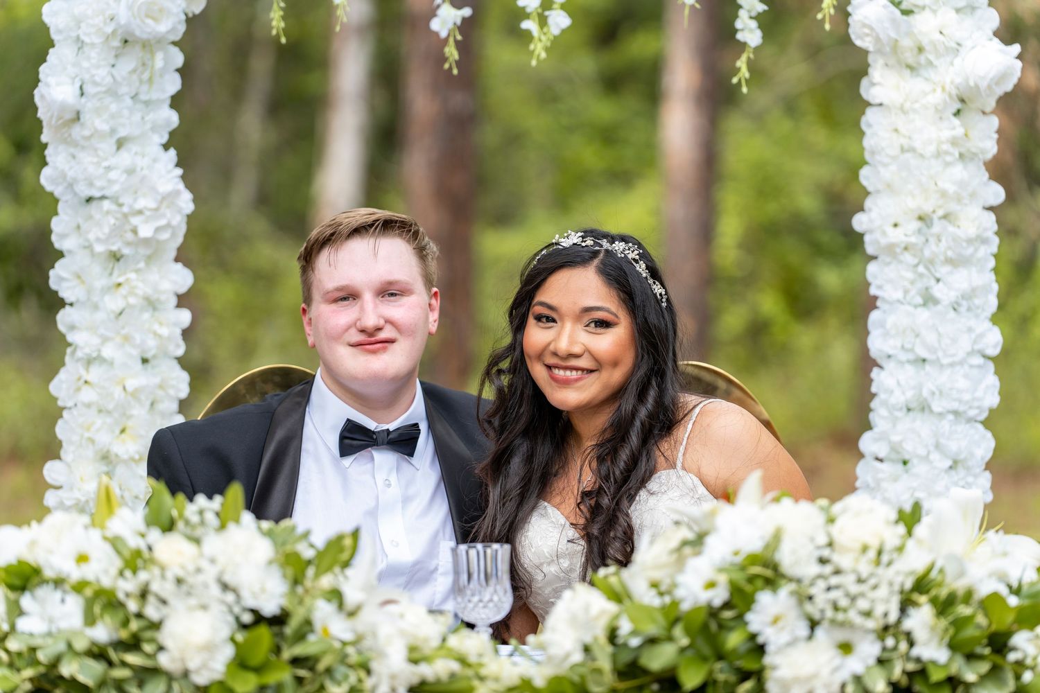 Smiling bride and groom seated at floral wedding reception table surrounded by white flowers and greenery outdoors.
