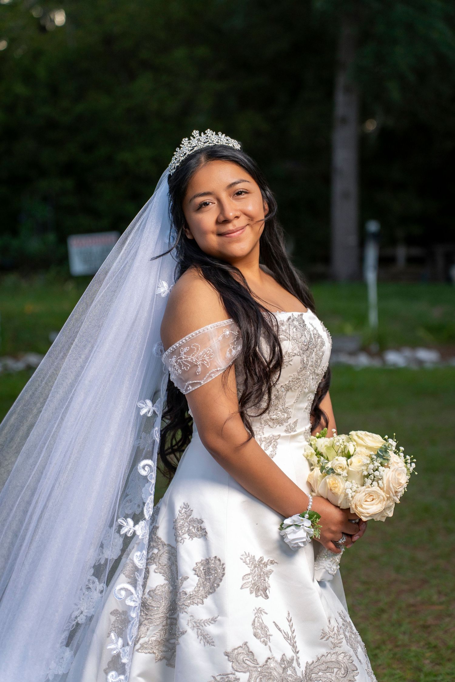 Smiling bride in white lace off-shoulder gown, tiara, and veil holds bouquet of white roses outdoors.