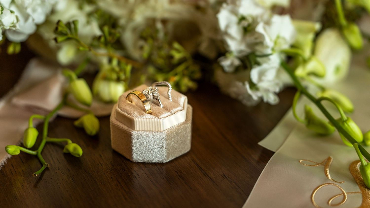 Gold wedding rings displayed in a hexagonal velvet ring box with white floral bouquet in background.