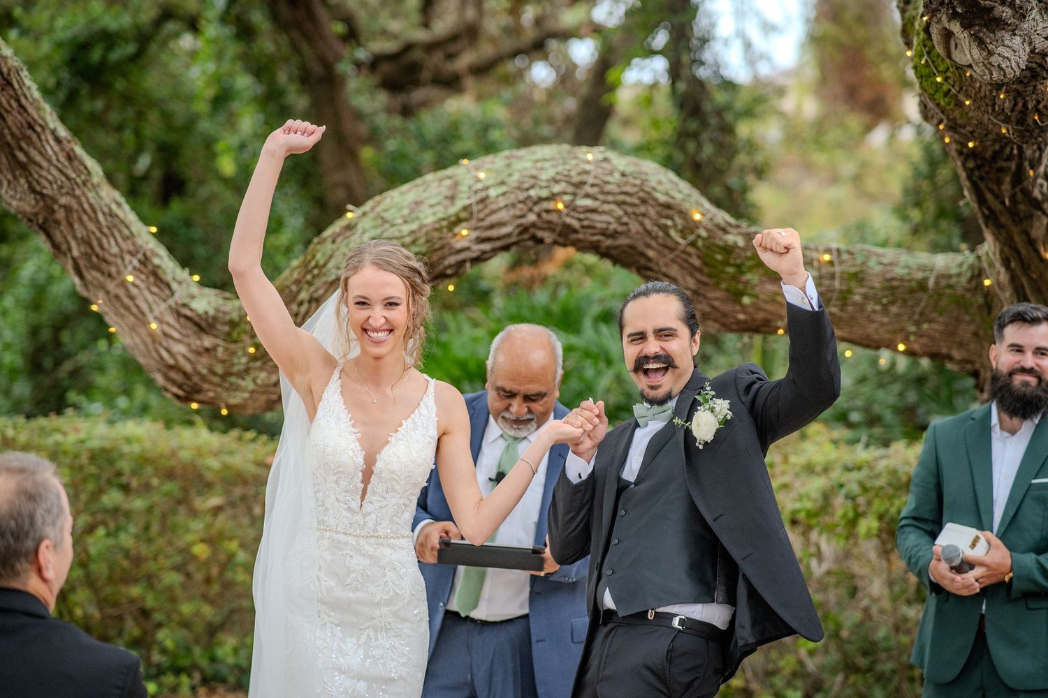 Wedding party joyfully celebrates on a lush green lawn with arms raised in celebration on a sunny day.