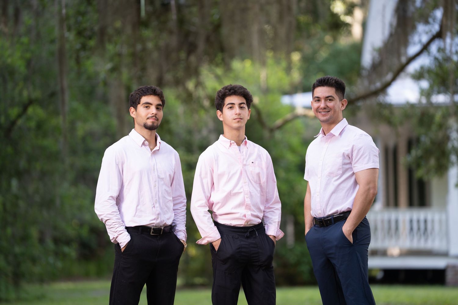 Three people in formal attire pose together outdoors with trees and a white building in the background.