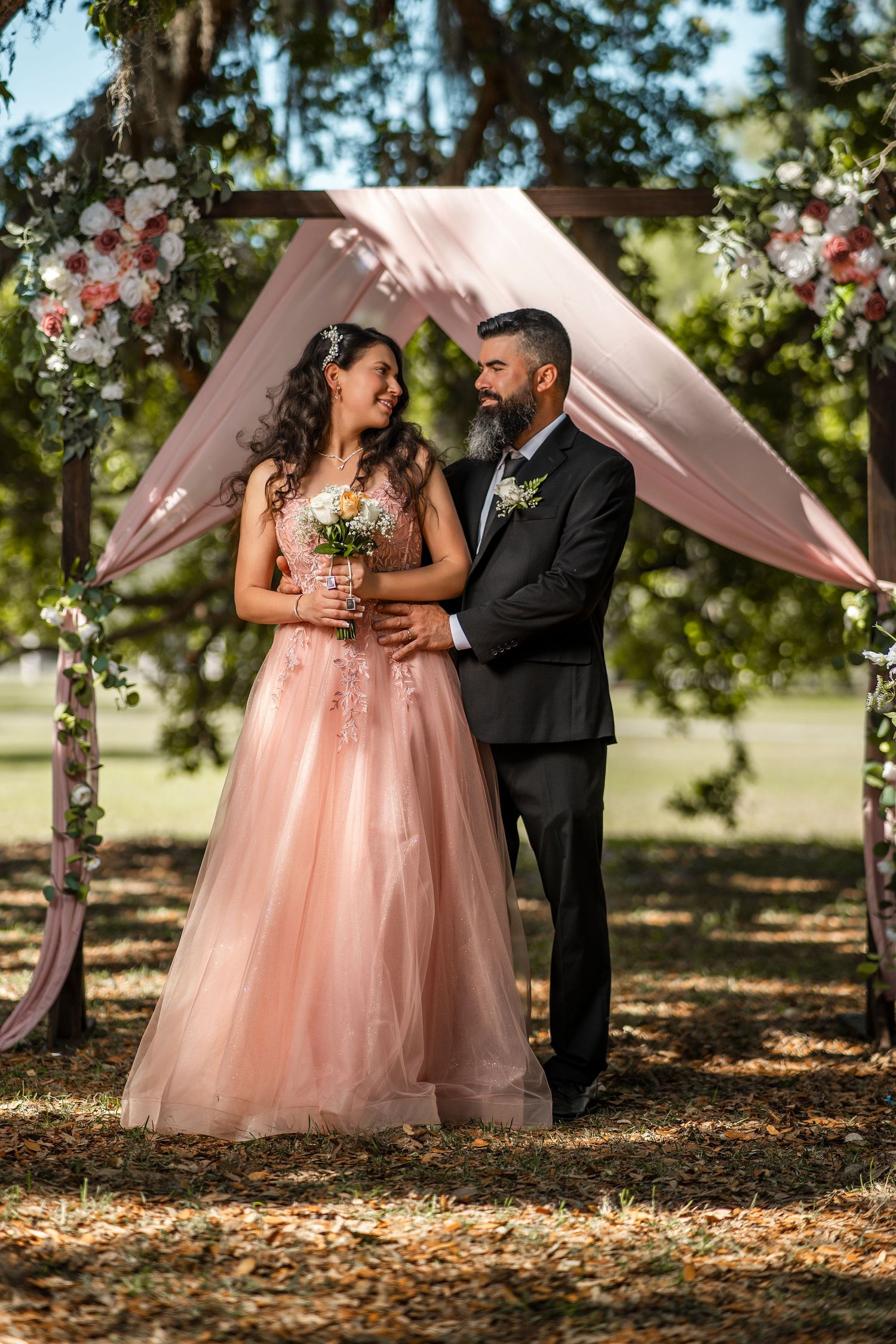 Couple in wedding attire standing under floral arch outdoors, bride in pink gown holding bouquet, groom in black suit.