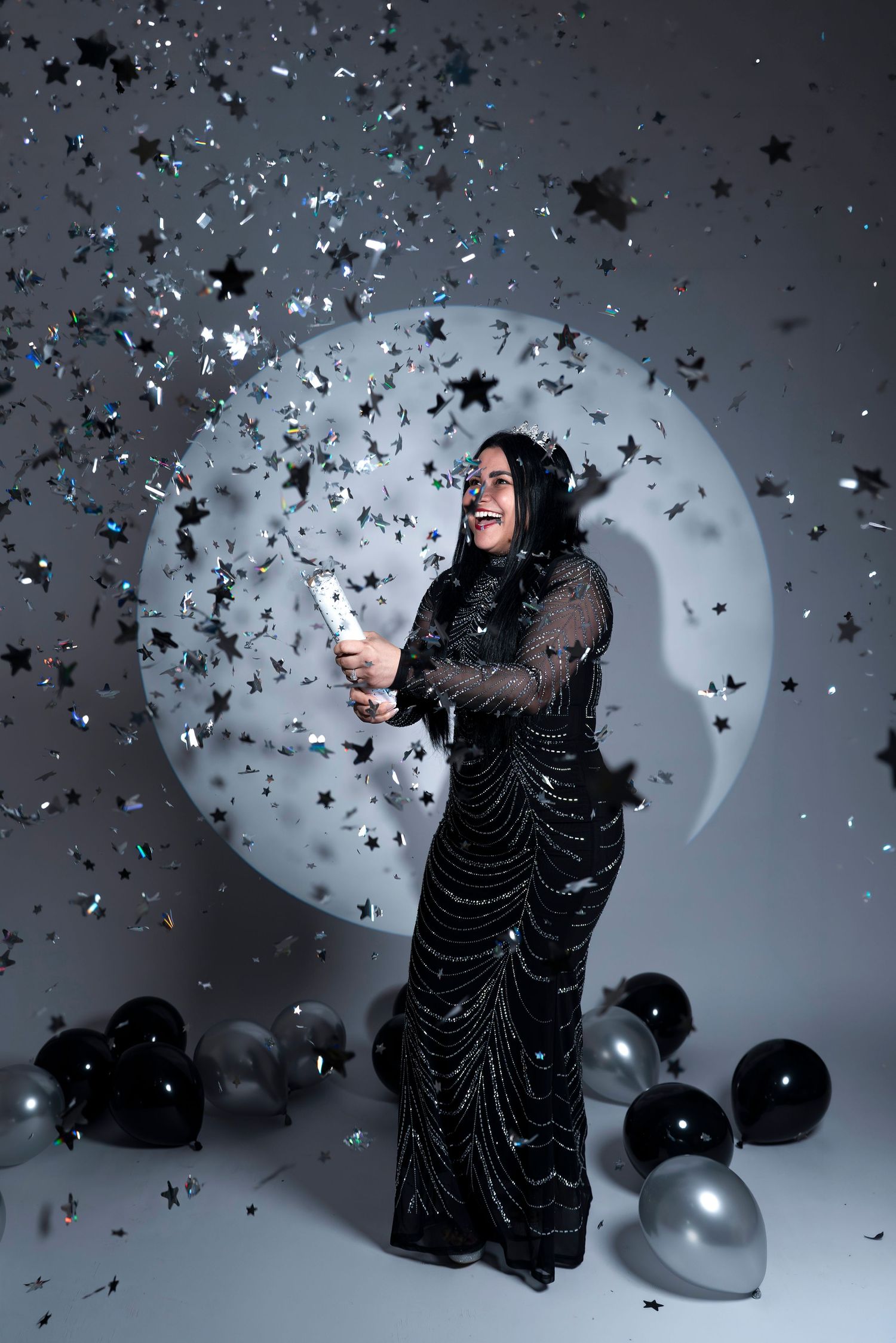 A person in a black sparkly dress celebrates with confetti and balloons against a circular light backdrop.