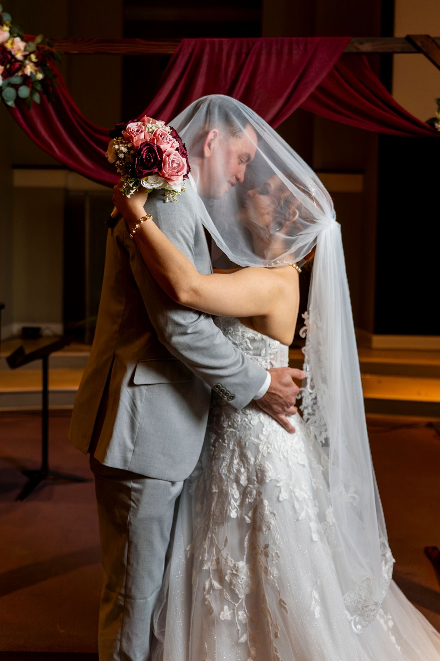 A romantic wedding couple embraces during their first dance in a warm-lit venue with flowing veil and floral details.