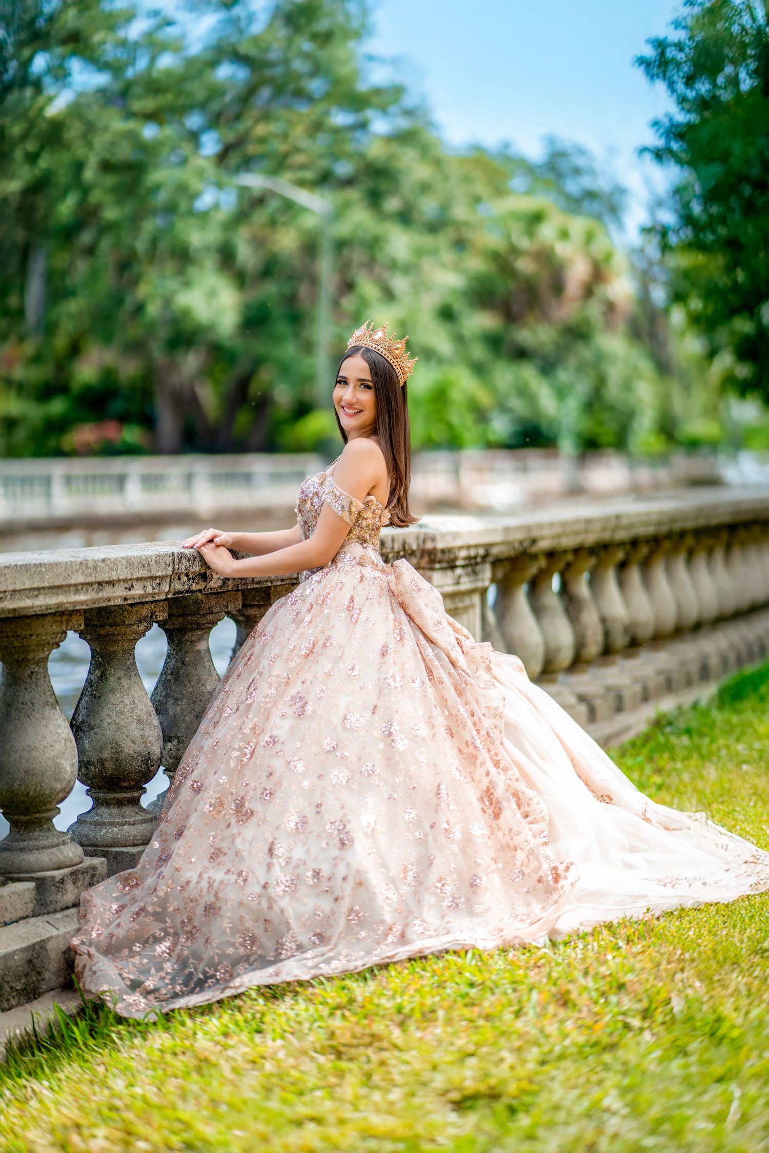Elegant ball gown in blush pink photographed against stone balustrade and lush garden backdrop.