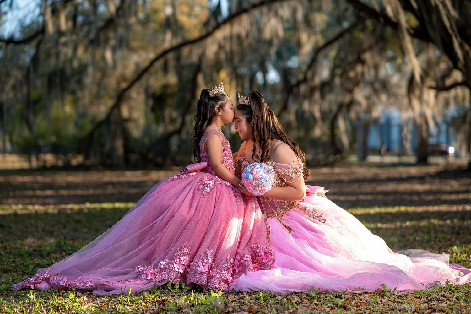 Two girls in matching pink ball gowns share a tender moment outdoors under moss-draped trees.