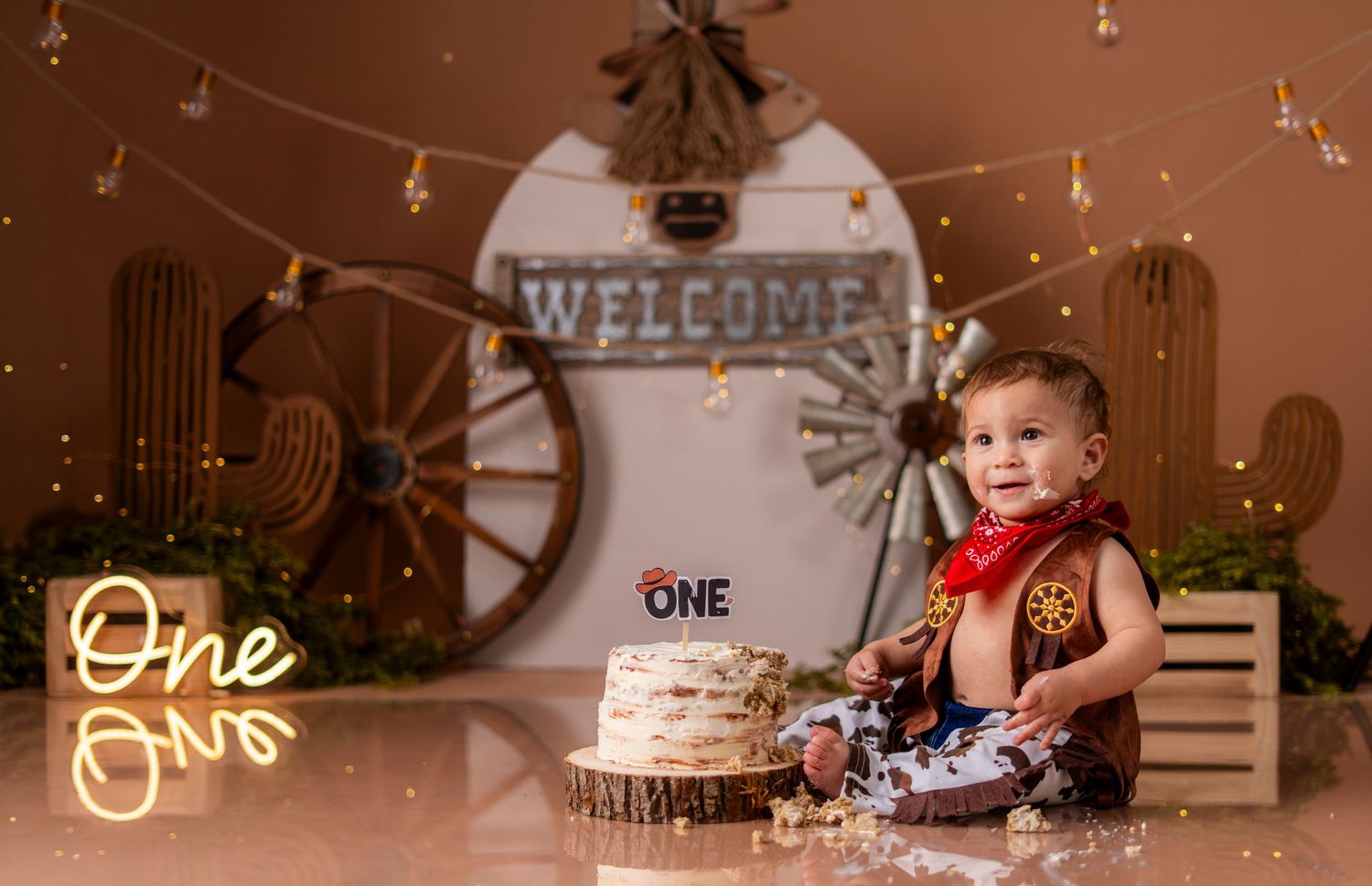 Rustic western themed first birthday setup with cake and string lights against wooden wagon wheel backdrop.