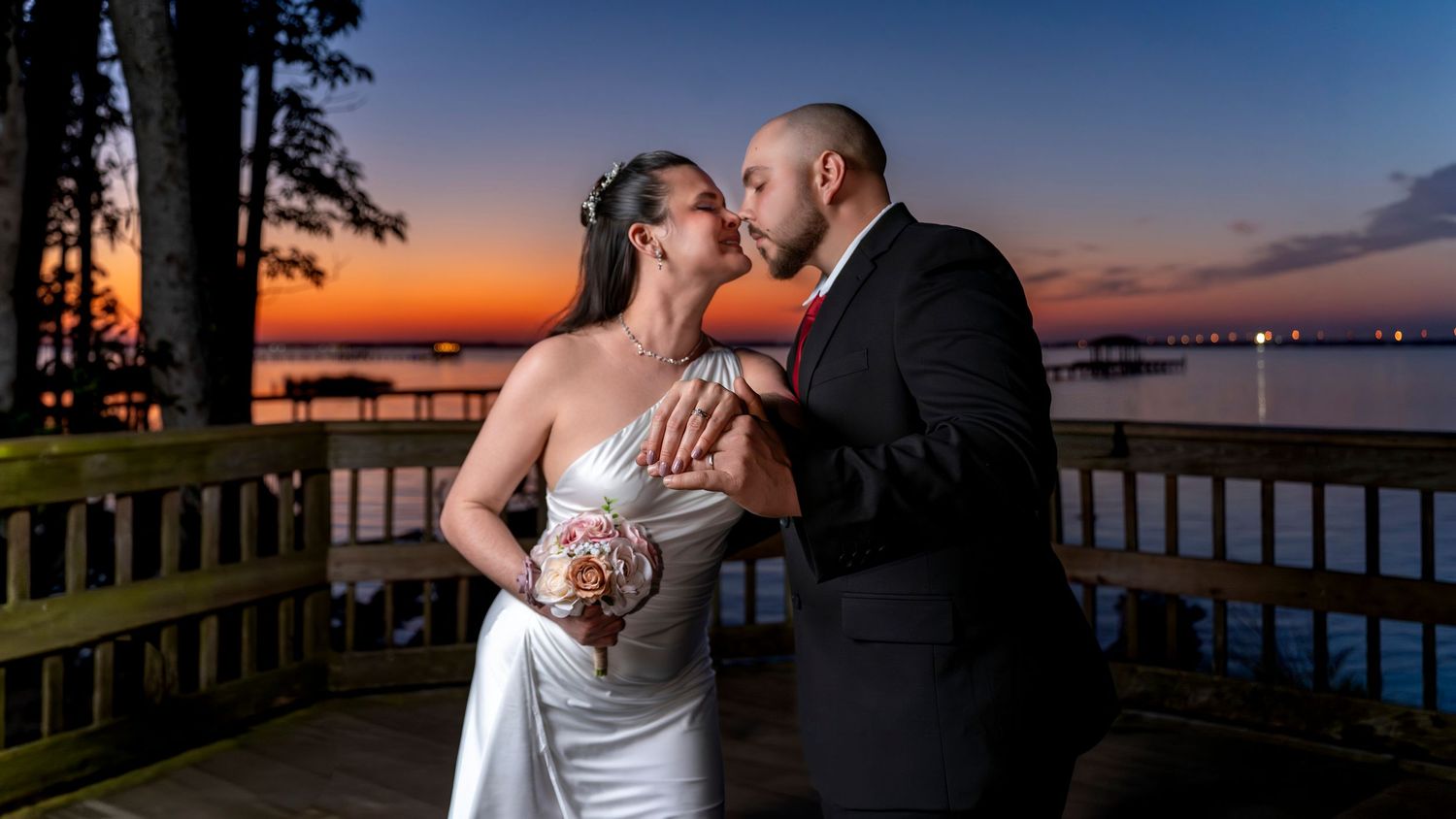 Bride and groom sharing a romantic kiss on a waterfront deck at sunset with colorful orange and blue sky backdrop.