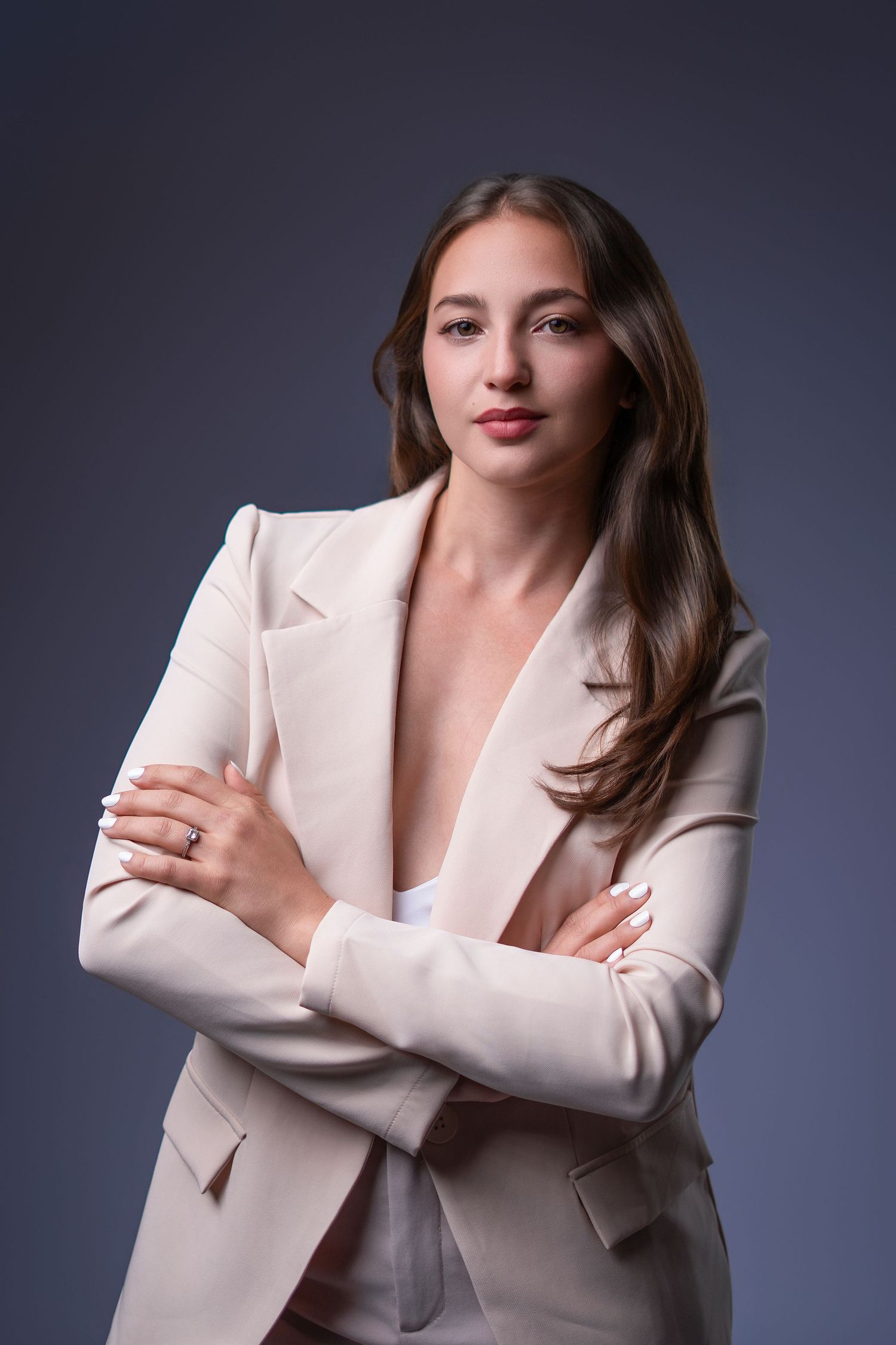 Young woman in a cream blazer with arms crossed, posing confidently against a dark gray studio background.