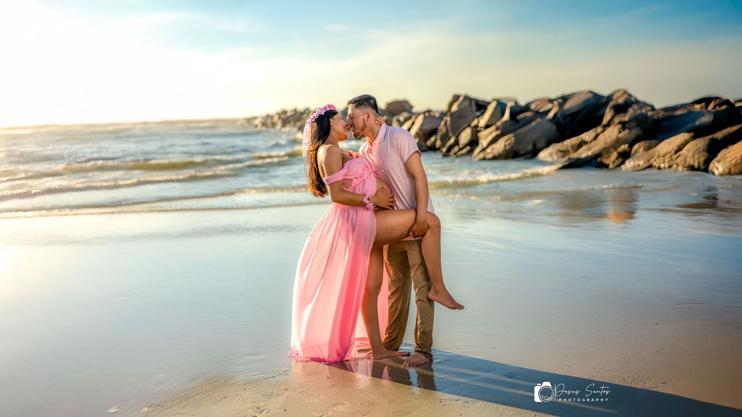 A couple embraces on a scenic beach with rocks and waves at sunset, person wearing a flowing pink dress.