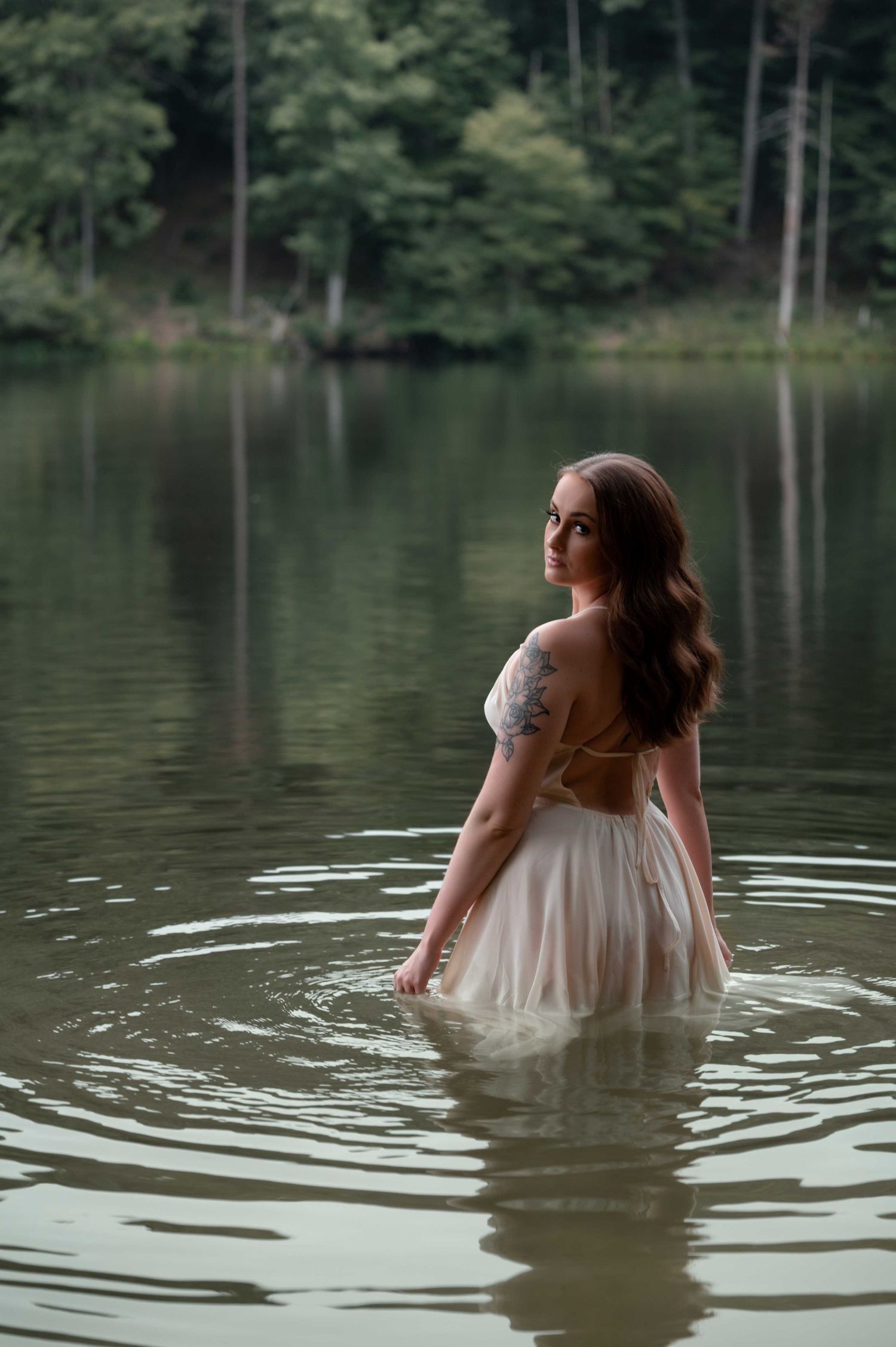 Romantic boudoir by lake – woman in dress standing in water, looking back with confidence.