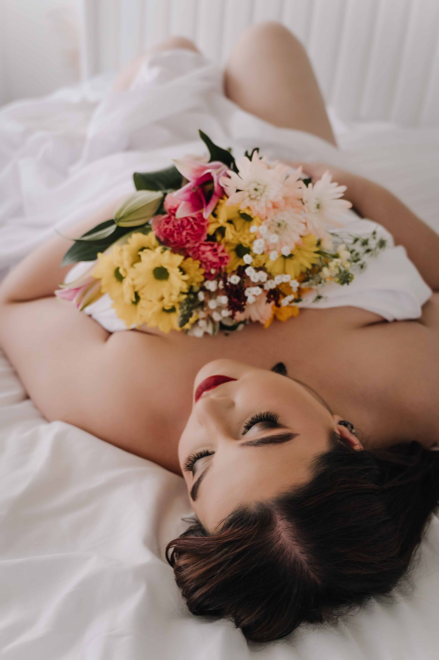 Boudoir photo of woman lying on white sheets with colorful flower bouquet across her chest