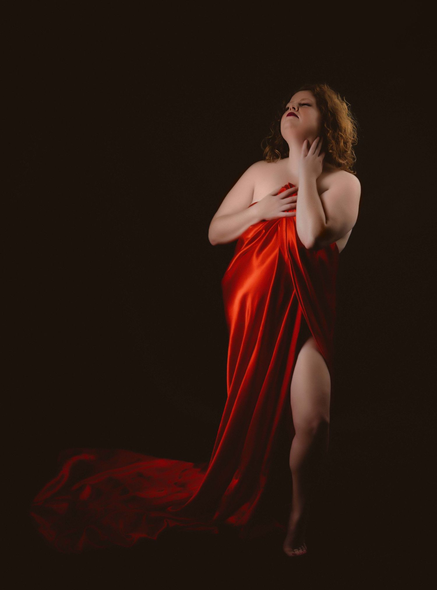 Curvy woman wrapped in rich red satin fabric poses against a dark background, fabric cascading to the floor as she tilts her head back