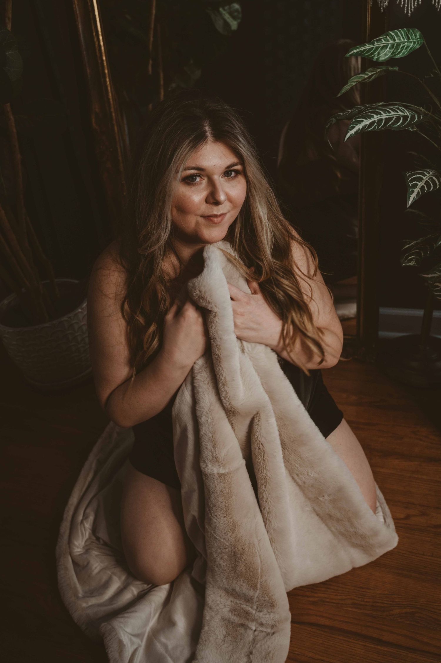 Woman with long hair kneeling on the floor holding a soft cream blanket to her chest, smiling warmly in a cozy, intimate scene.