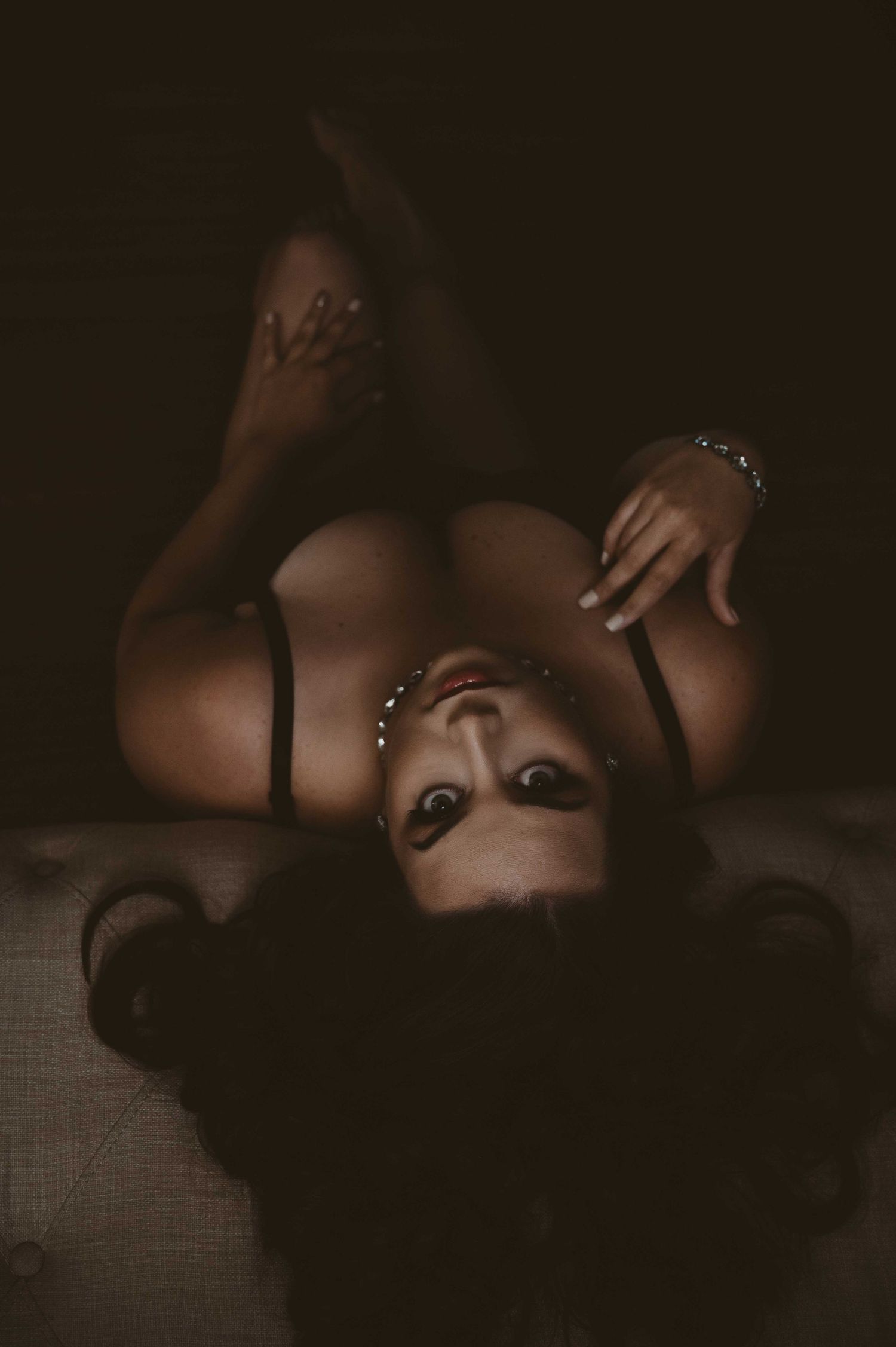 Woman lying upside down on a bench in a dark moody setting, wearing black lingerie and jewelry, looking directly into the camera.