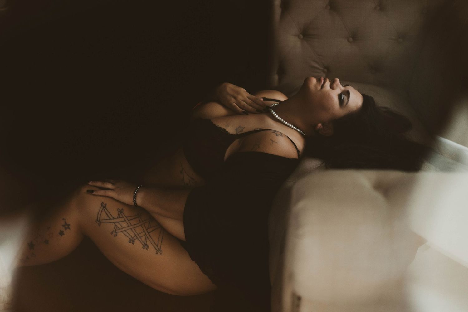Dark and moody boudoir pose – Tattooed woman in black lingerie with pearls, reclining by a tufted chair.