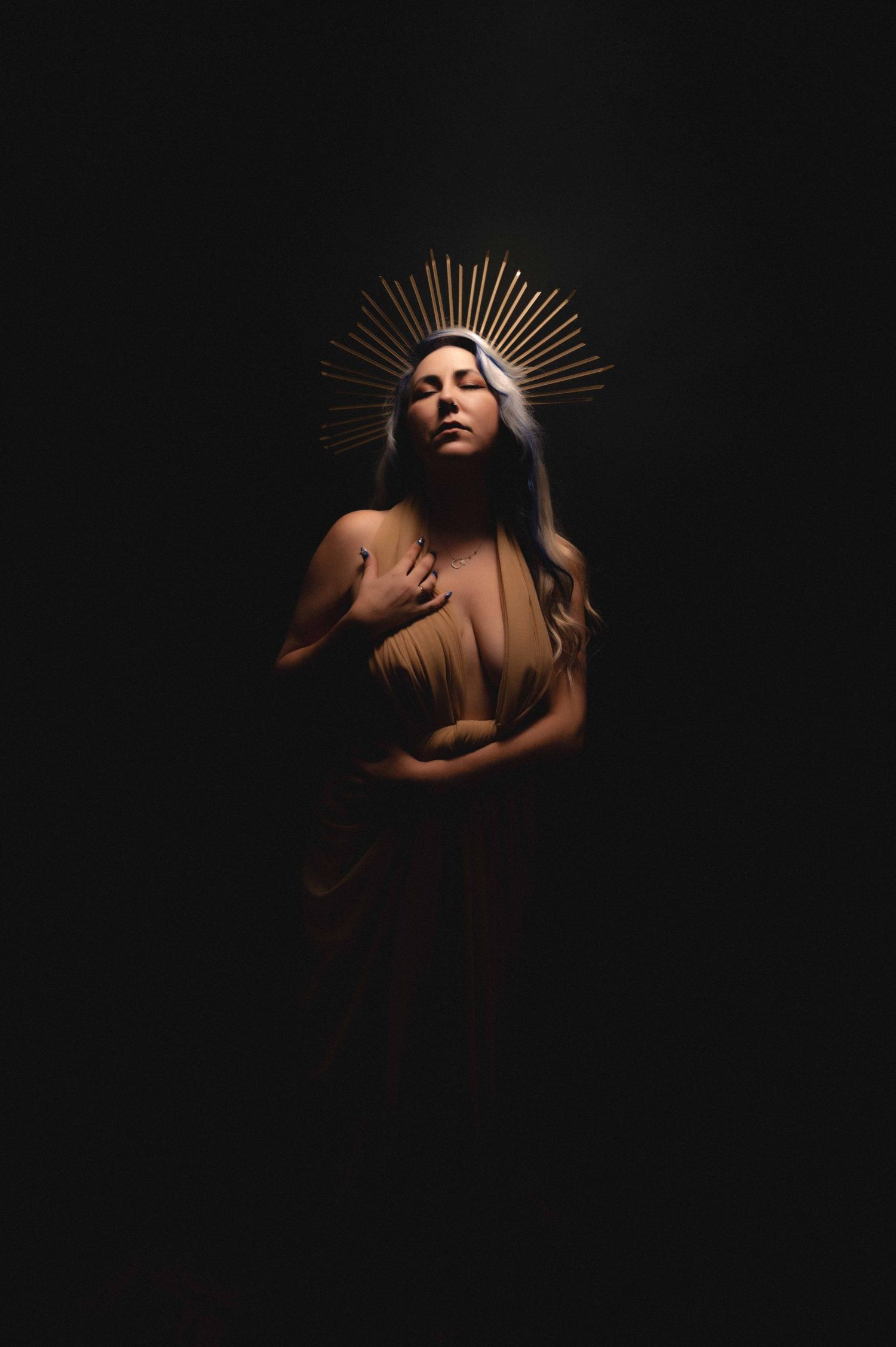 Woman in golden draping stands illuminated against a dark backdrop, wearing a radiant crown headpiece, hands resting on her chest