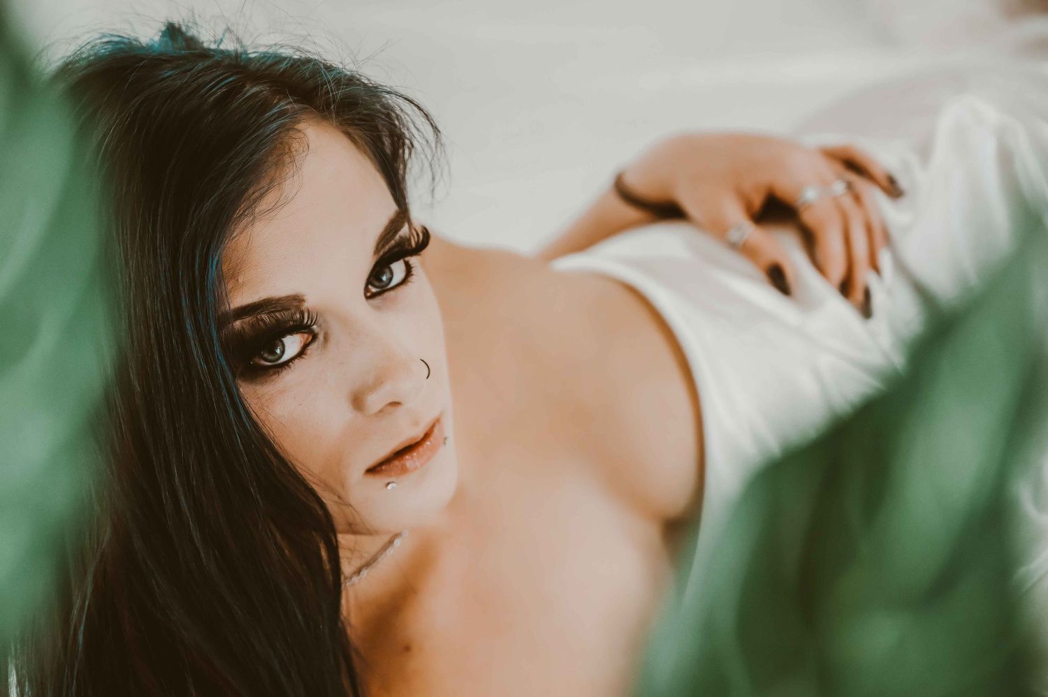 Dark-haired woman on white sheets, close-up gaze framed by blurred greenery.