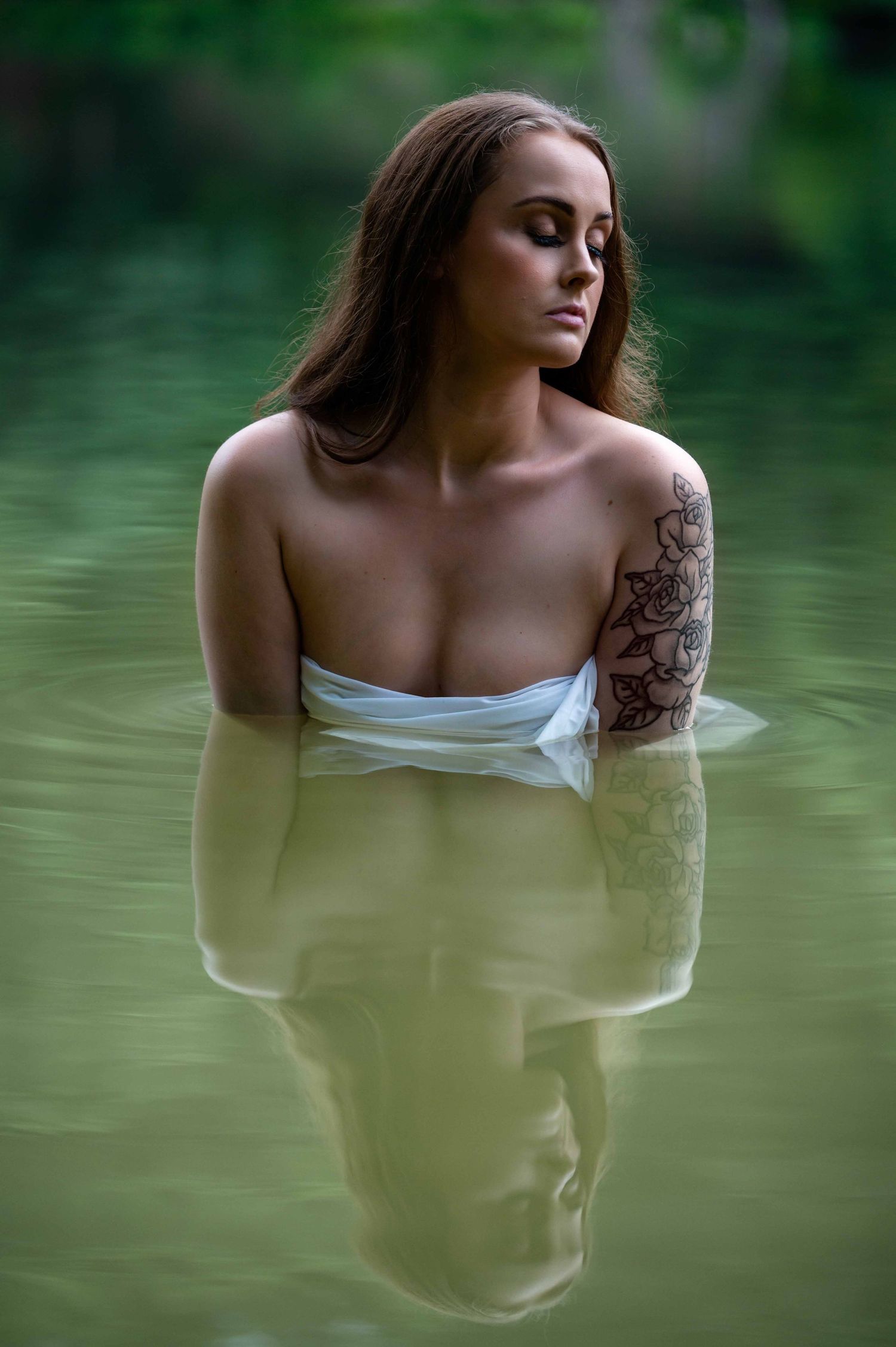 Outdoor boudoir shoot of a woman wrapped in white fabric, sitting in a lake, serene intimate portrait photography.