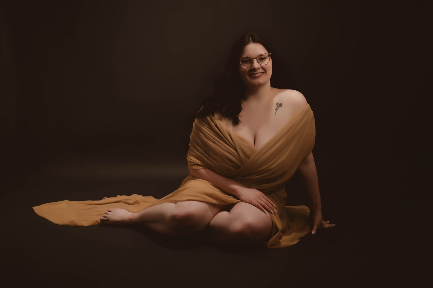 Woman with dark wavy hair and glasses sits on the floor against a dark backdrop, wrapped in a flowing mustard-colored drape, smiling warmly.