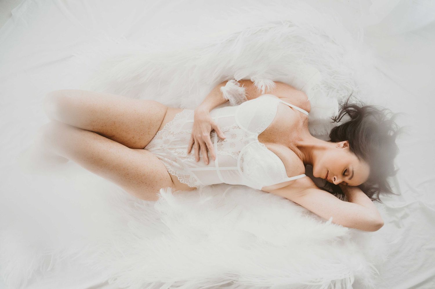 Woman in white lace lingerie and angel wings lying on a bed surrounded by fluffy white feathers.