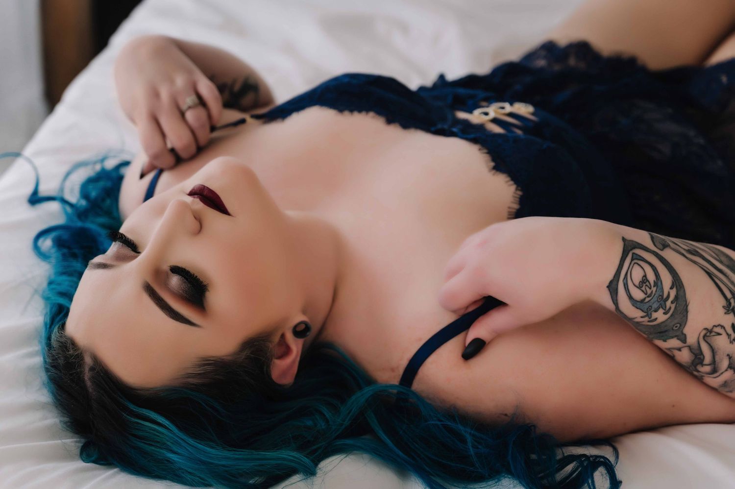 Woman with bright blue hair lying on a bed in navy lace lingerie, pulling on her strap with eyes closed in a sensual pose.