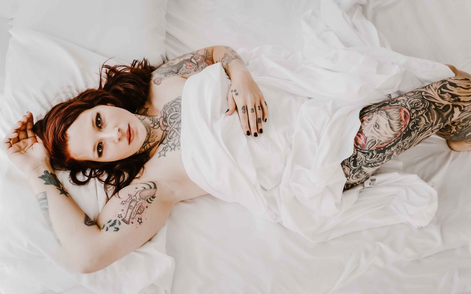 Boudoir photography Ashland KY – tattooed woman draped in white sheets, confident and powerful.