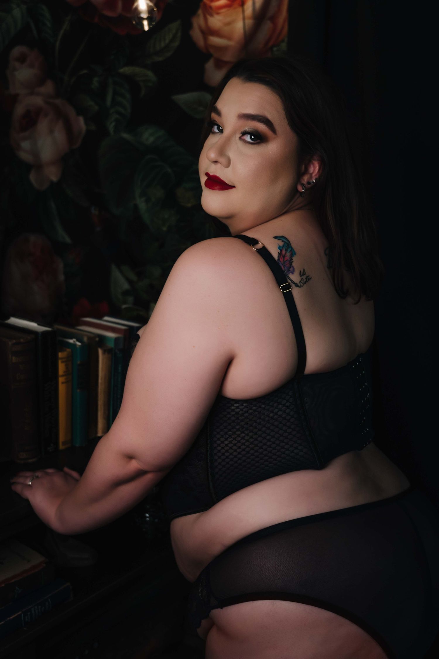 Confident boudoir portrait of woman with bold makeup and red lips in black lingerie, looking over shoulder by bookshelf