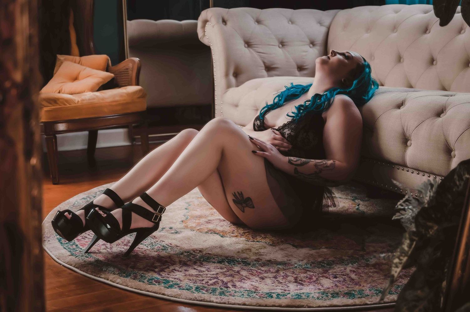 Woman with blue hair in black lace lingerie sitting on the floor against a tufted sofa, head tilted back in a relaxed, powerful pose.
