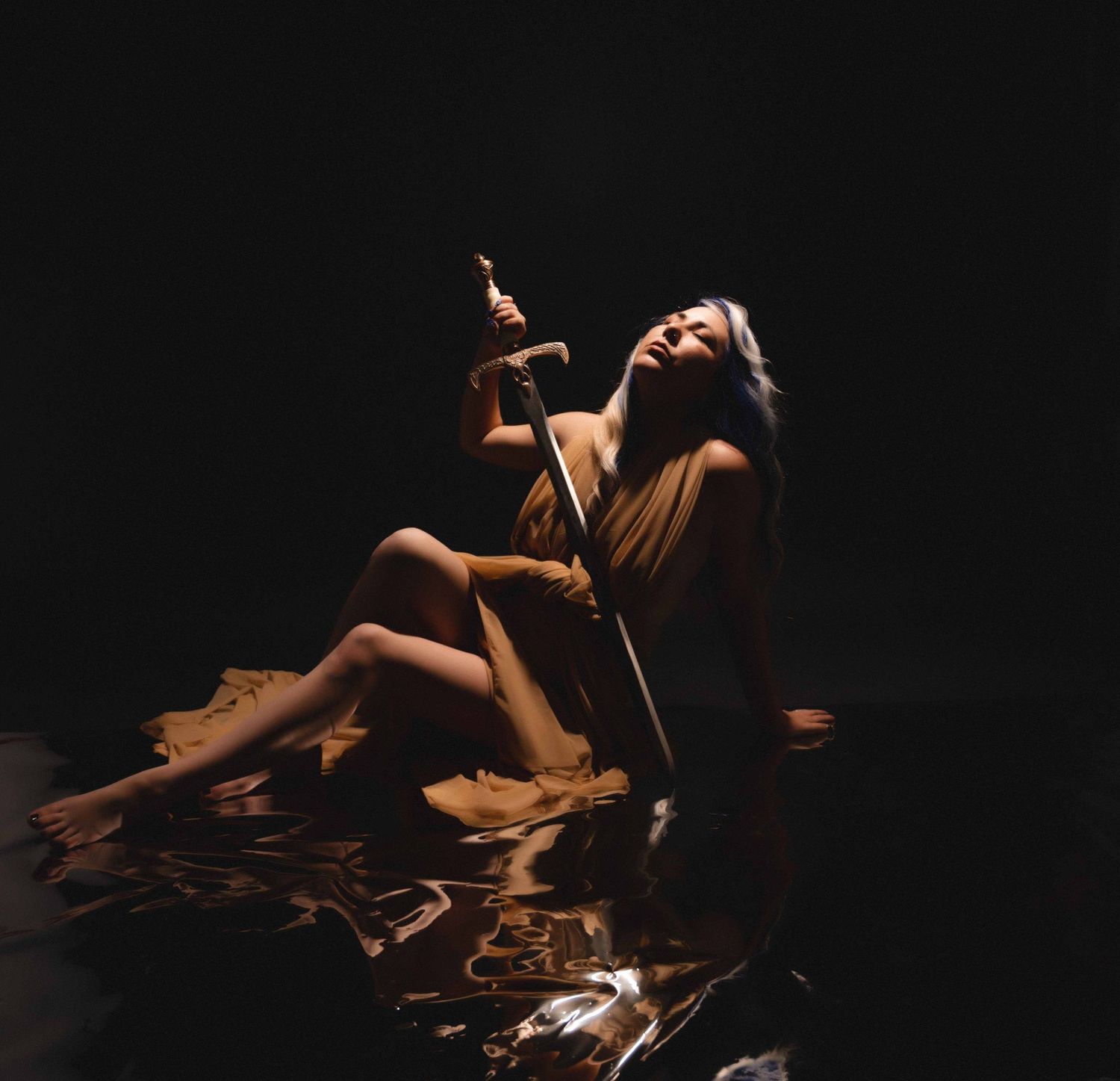 Woman in golden gown holding a sword, seated on reflective floor with dramatic low light.