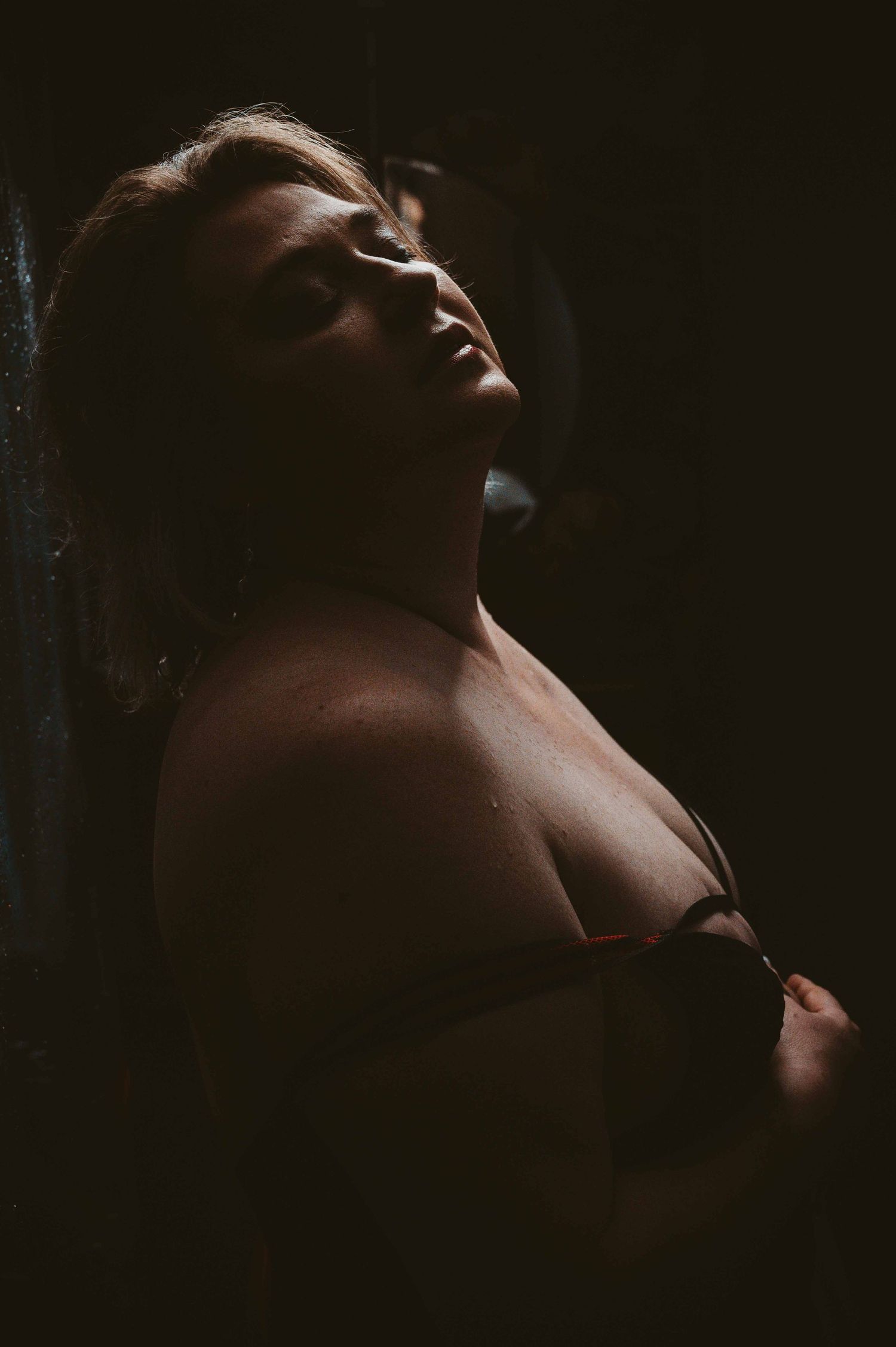 Moody boudoir photo in low light, woman in lingerie tilts her head back, artistic intimate photography in Kentucky.