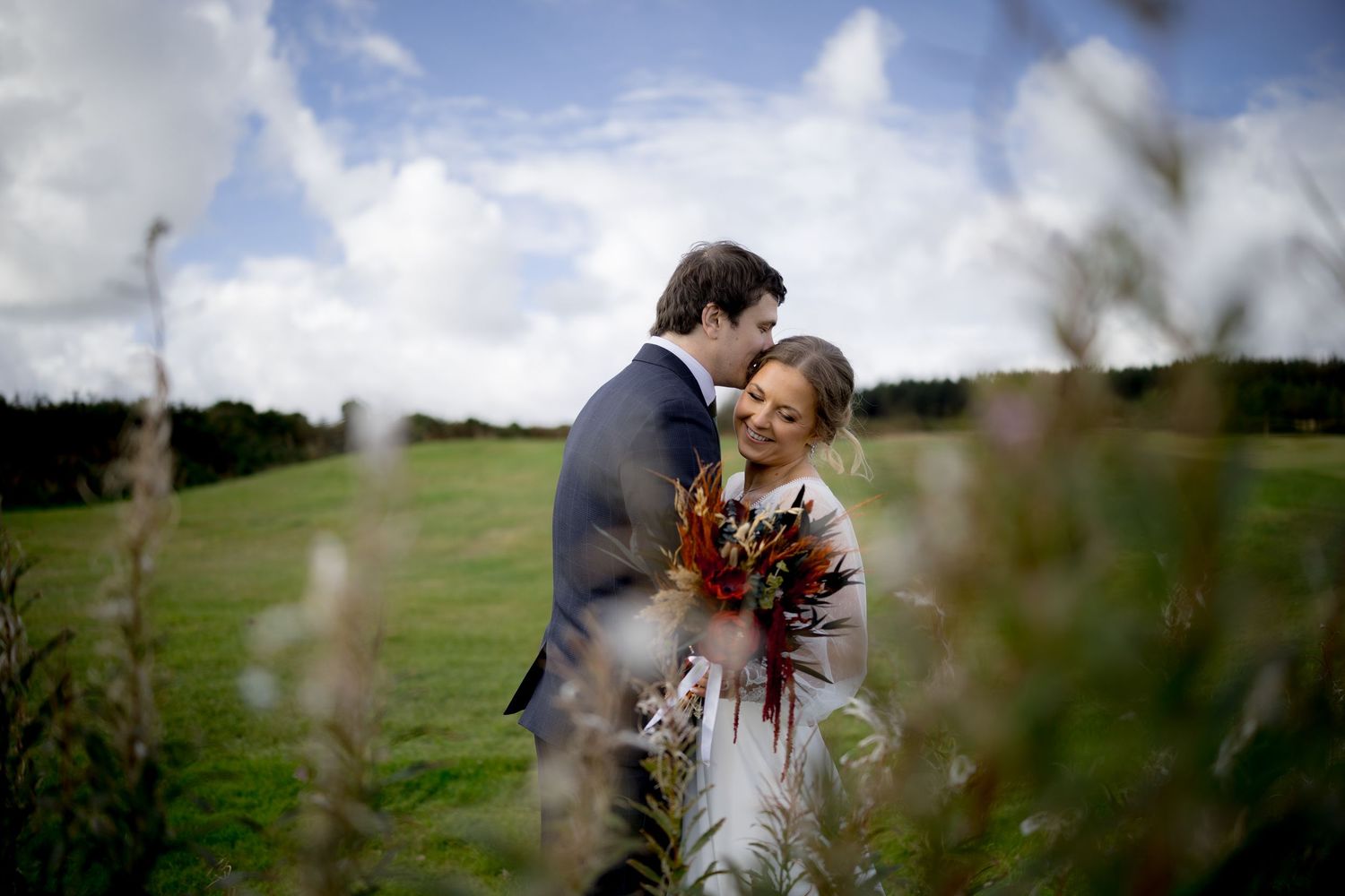 Bride and groom embrace in a lush green field, surrounded by wildflowers, under a partly cloudy sky on their wedding day.