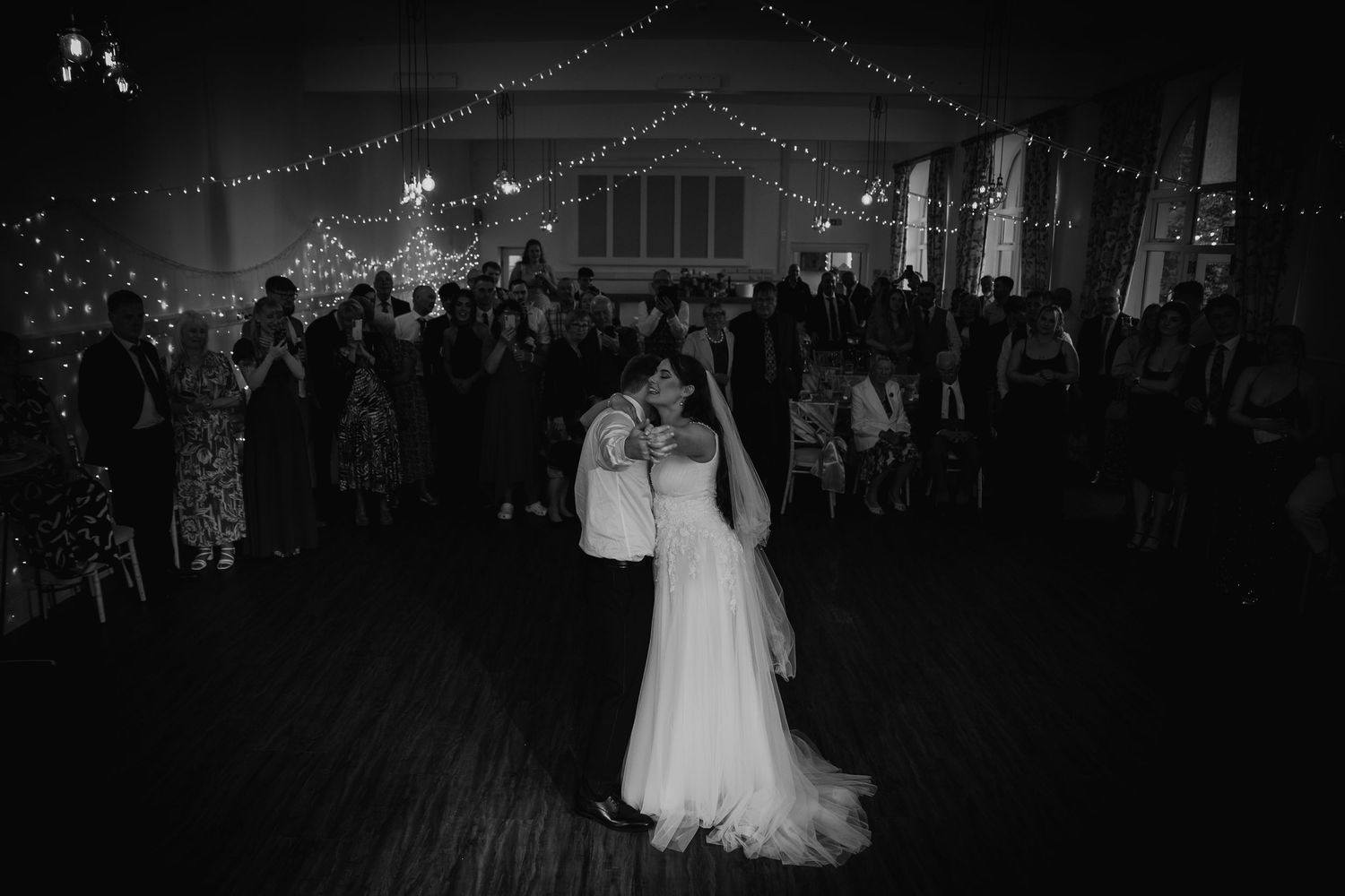 Black and white photo of bride and groom sharing first dance at wedding reception with fairy lights.