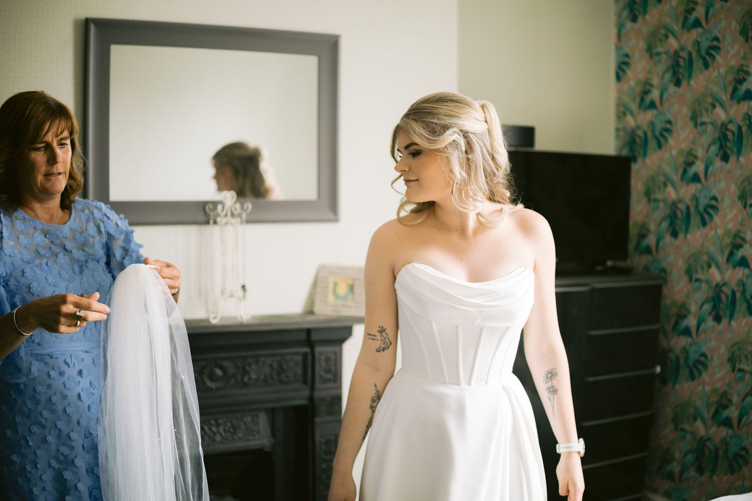 Bride in white strapless gown being helped by woman in blue dress in elegant room with mirror.
