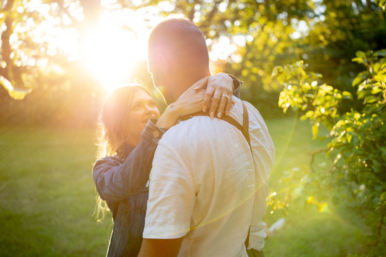 Couple embracing in a sunlit park with golden hour light and lush green trees in the background.