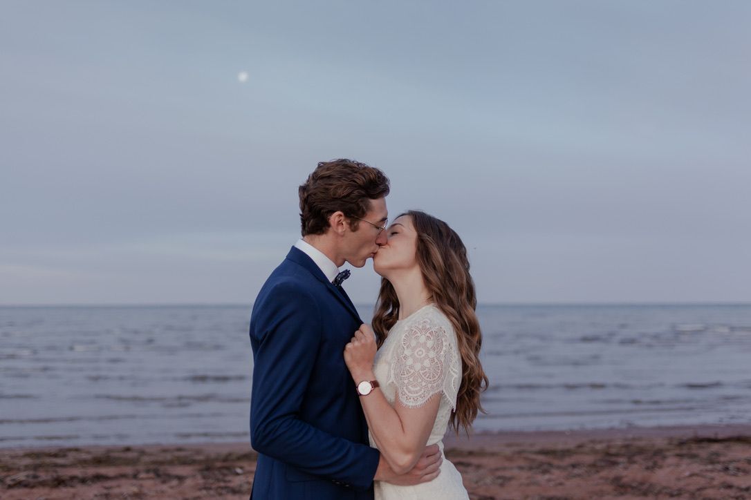 Elopement portraits of a couple kissing, at sunrise, on an Atlantic Canada beach. The moon can be seen in the background