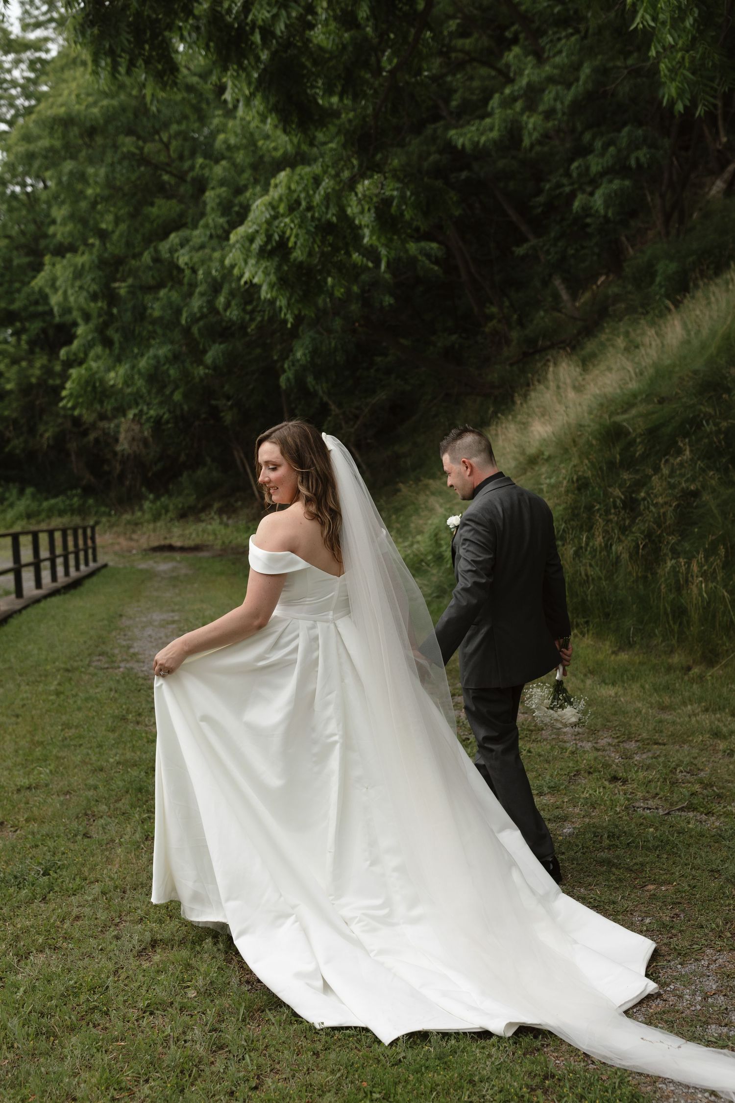 Bride in white off-shoulder gown with long train walks with groom in dark suit outdoors on green path.