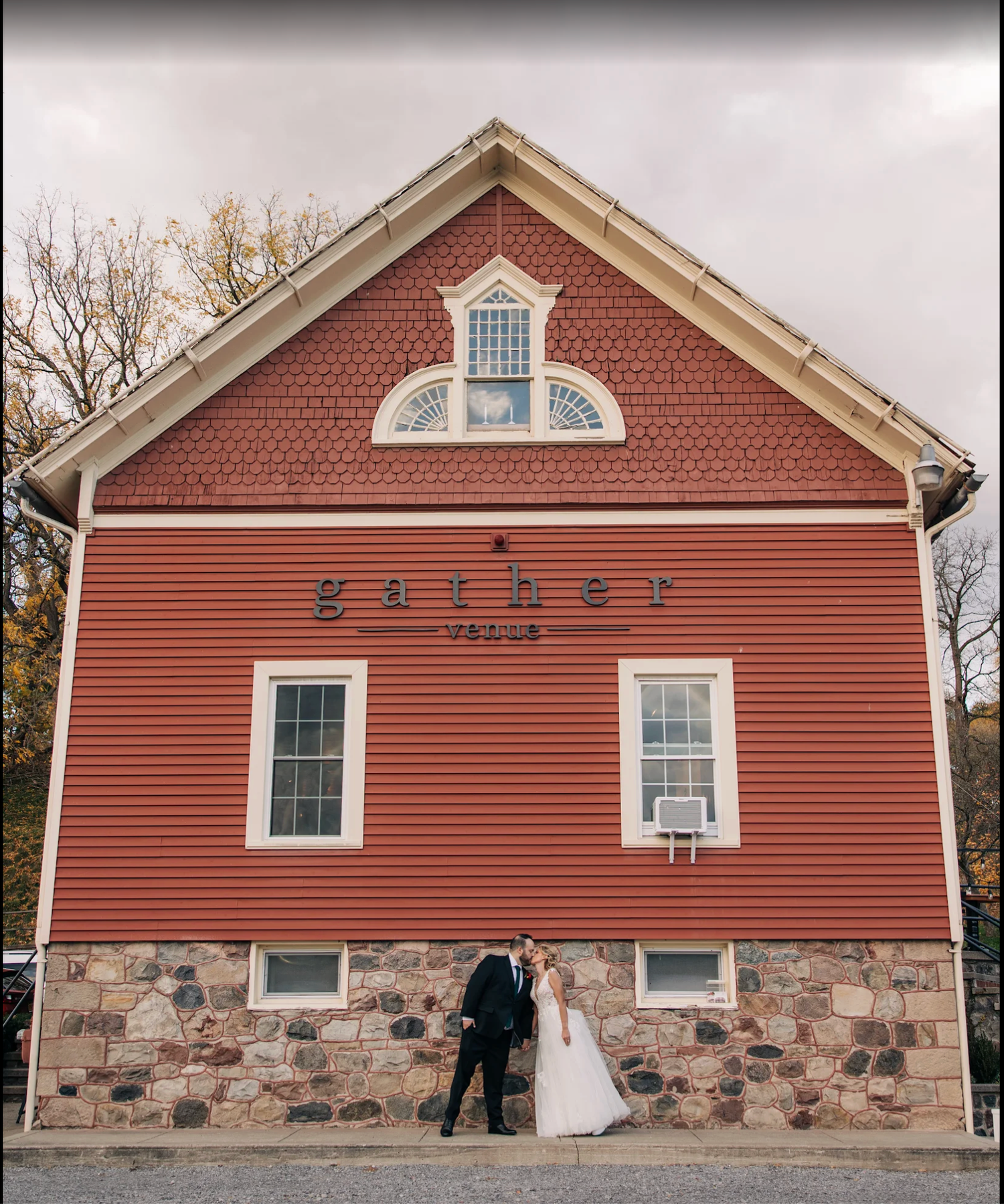 Bride and groom kissing in front of red Gather Venue barn with stone foundation on their wedding day.