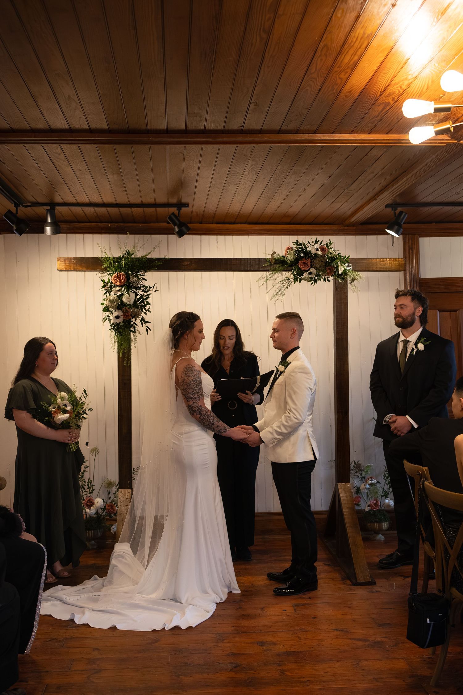 Bride and groom exchange vows at rustic indoor wedding ceremony with floral arch, officiant, bridesmaid, and groomsman.