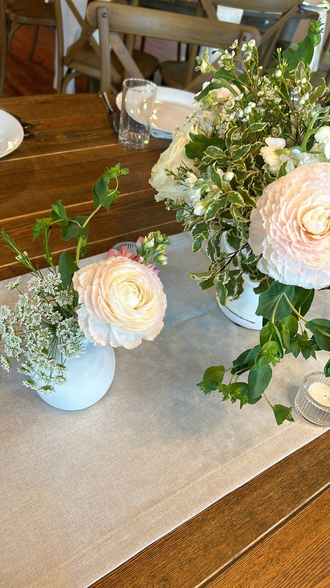 Elegant table centerpiece with blush pink dahlias and ranunculus in white vases with lush greenery on a rustic wood table.