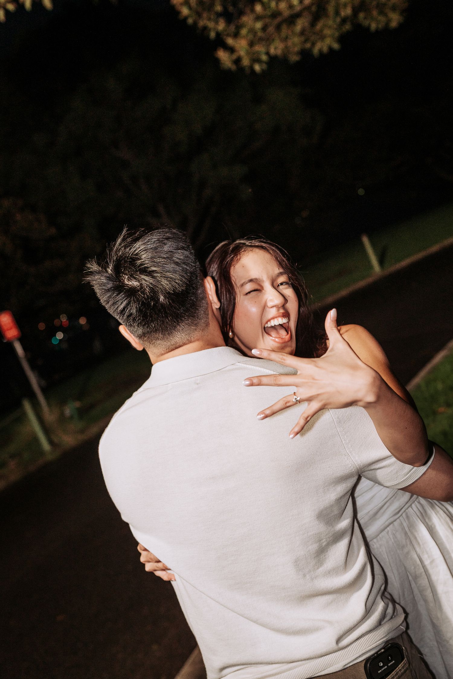 Joyful couple embracing outdoors at night, woman laughing with engagement ring visible on her hand.