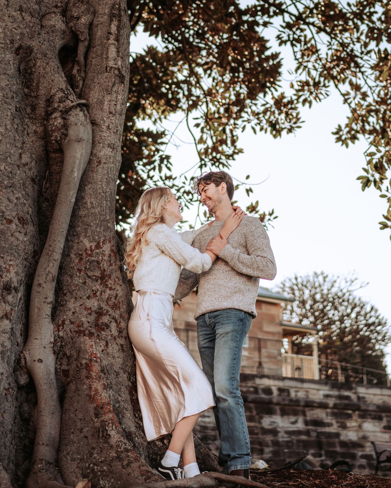 A romantic couple poses together in an intimate moment against a large tree trunk in a scenic outdoor setting.