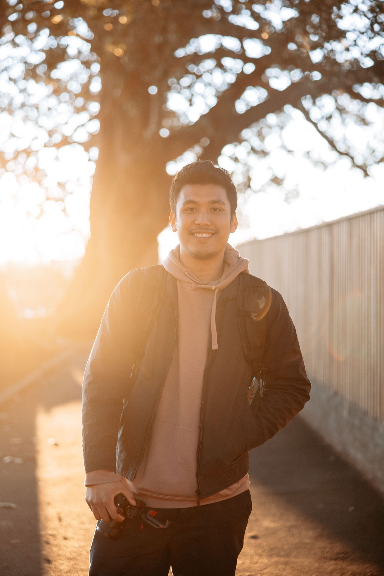 A casually dressed person stands smiling in warm sunset light near a white fence.