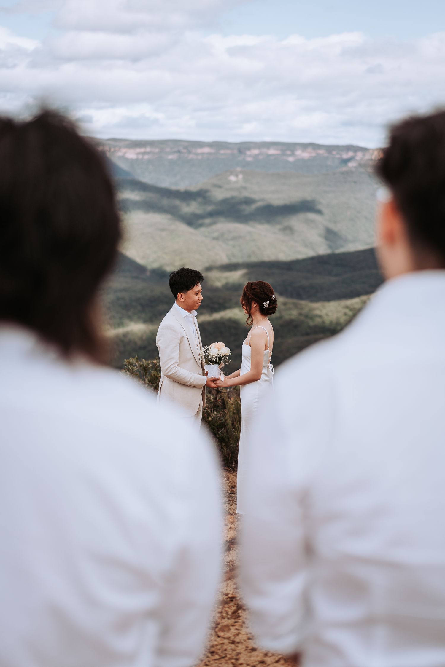 A couple in white wedding attire exchanges vows on a mountaintop with a dramatic valley landscape in the background.